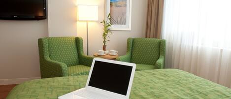 A hotel room with a bed, two green armchairs, a laptop on the bed, a TV on the wall, a lamp, a framed picture, and a window with curtains.