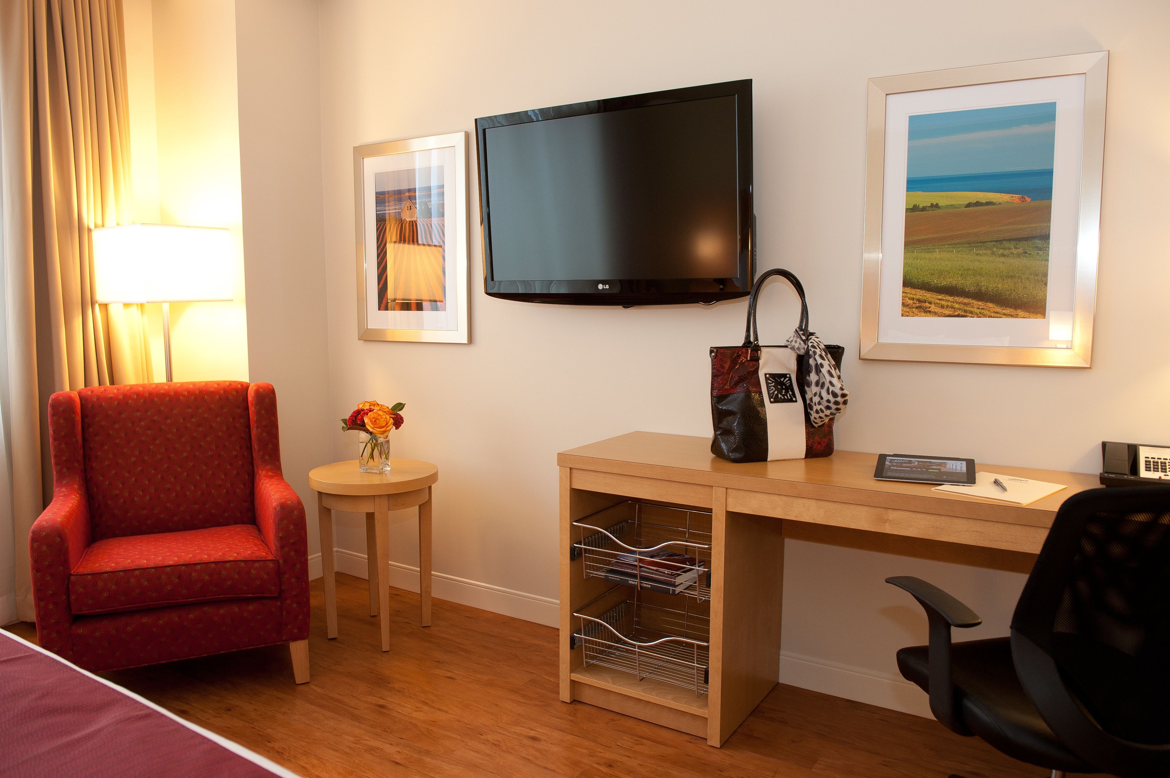 A hotel room with a red armchair, a desk with a computer, a TV, and a framed landscape picture on the wall.