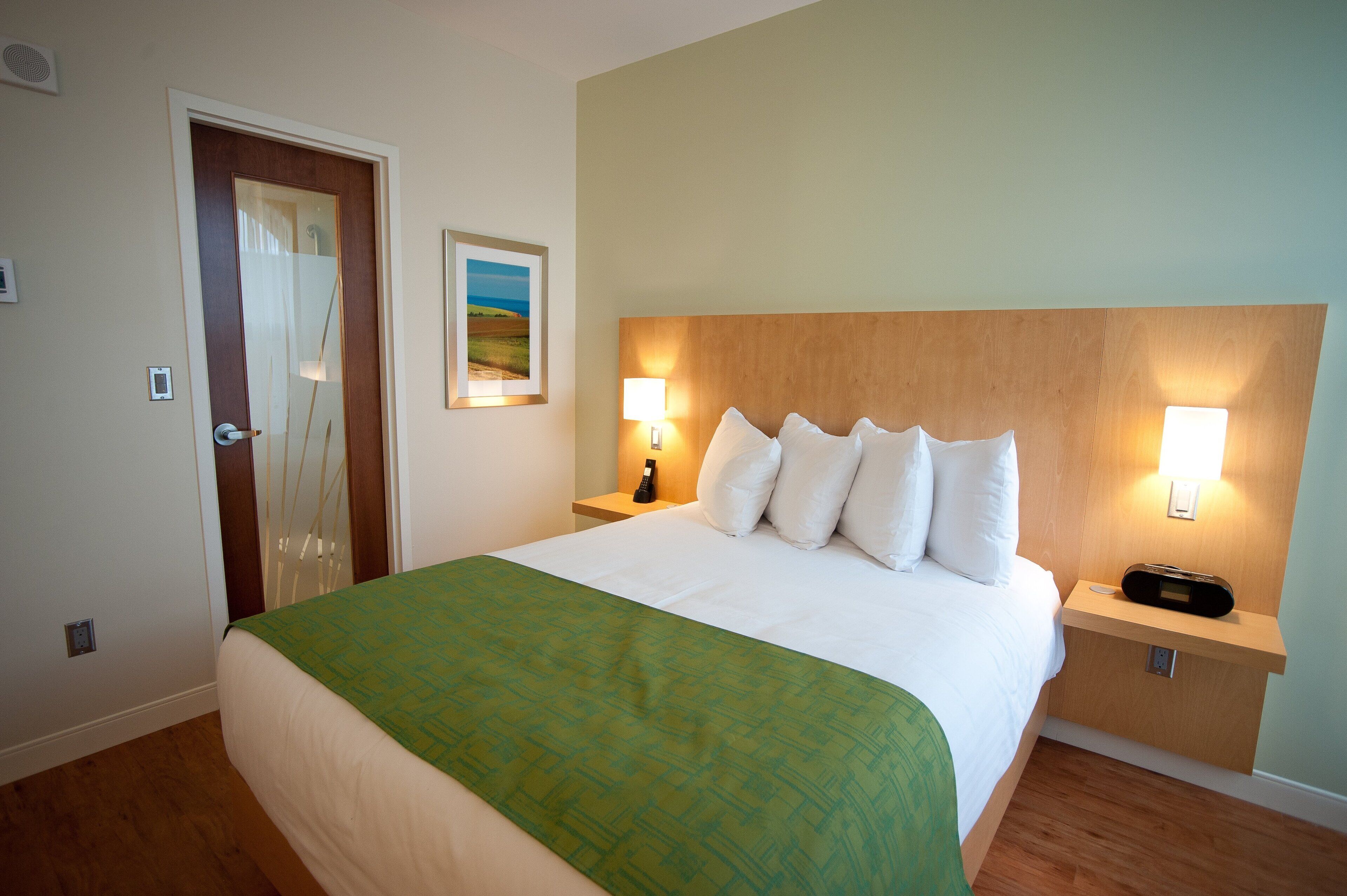 A hotel room with a large bed, a green bedspread, two bedside tables with lamps, a wooden headboard, and a door with a view of the outdoors.