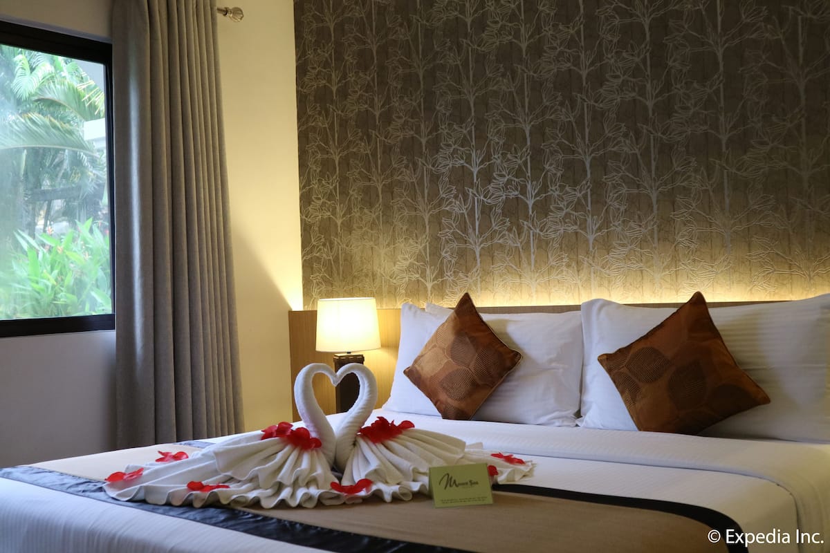 A hotel room with a neatly made bed, a heart-shaped towel arrangement, and a view of greenery through the window.