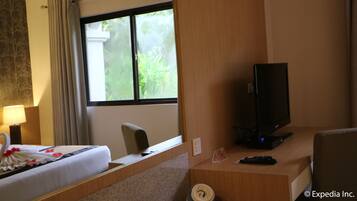 A hotel room with a bed, a desk with a television, a chair, a lamp, and a window with curtains.
