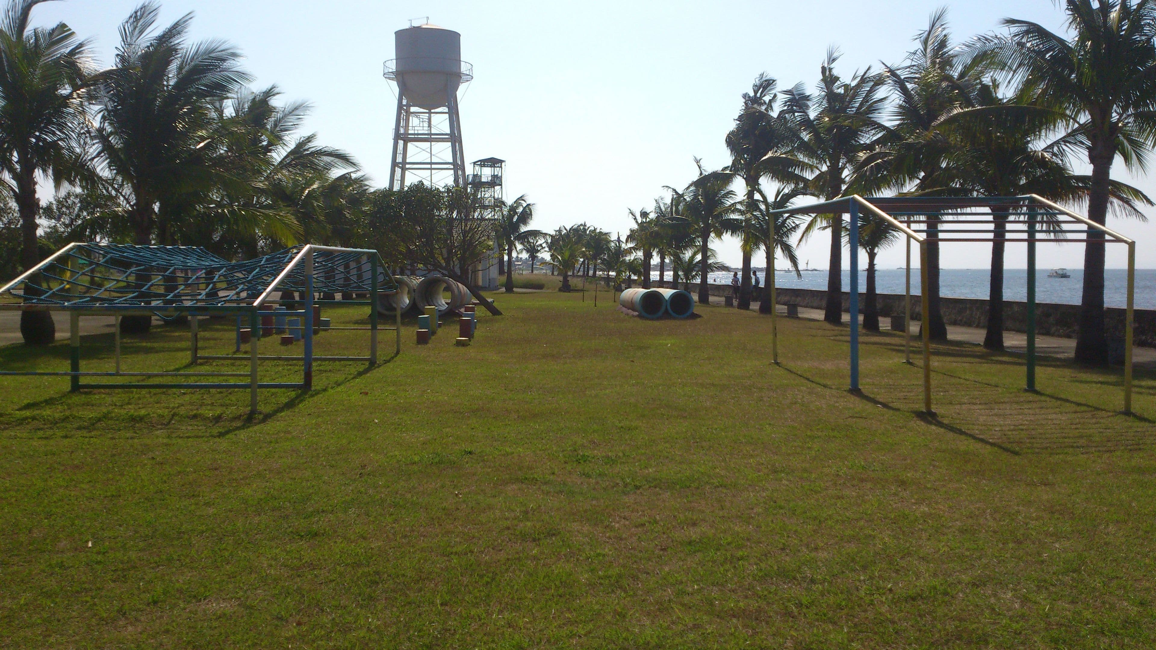 A grassy park with playground equipment, palm trees, and a water tower in the background.