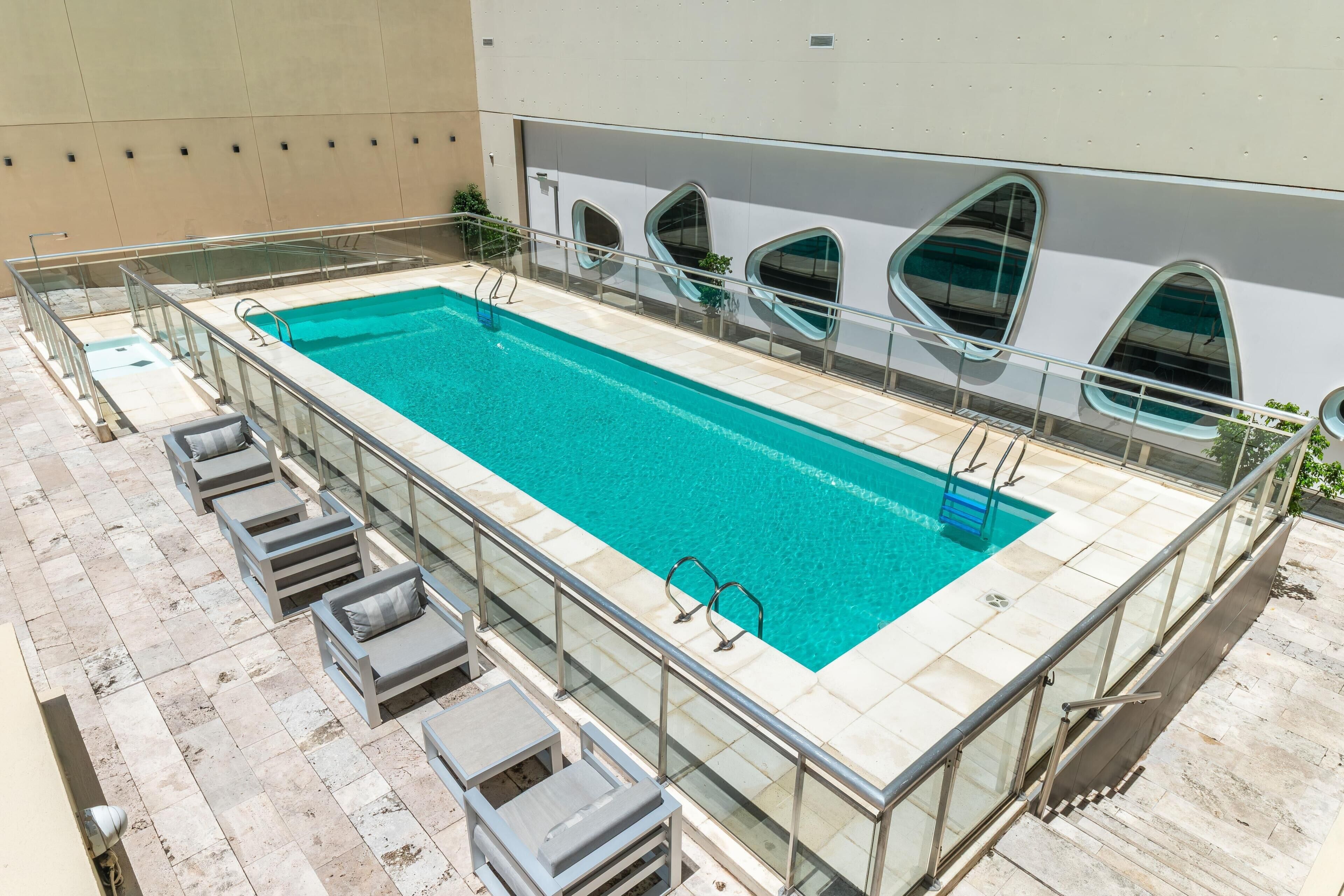 A rooftop swimming pool with glass railings, lounge chairs, and a modern building with unique window shapes.