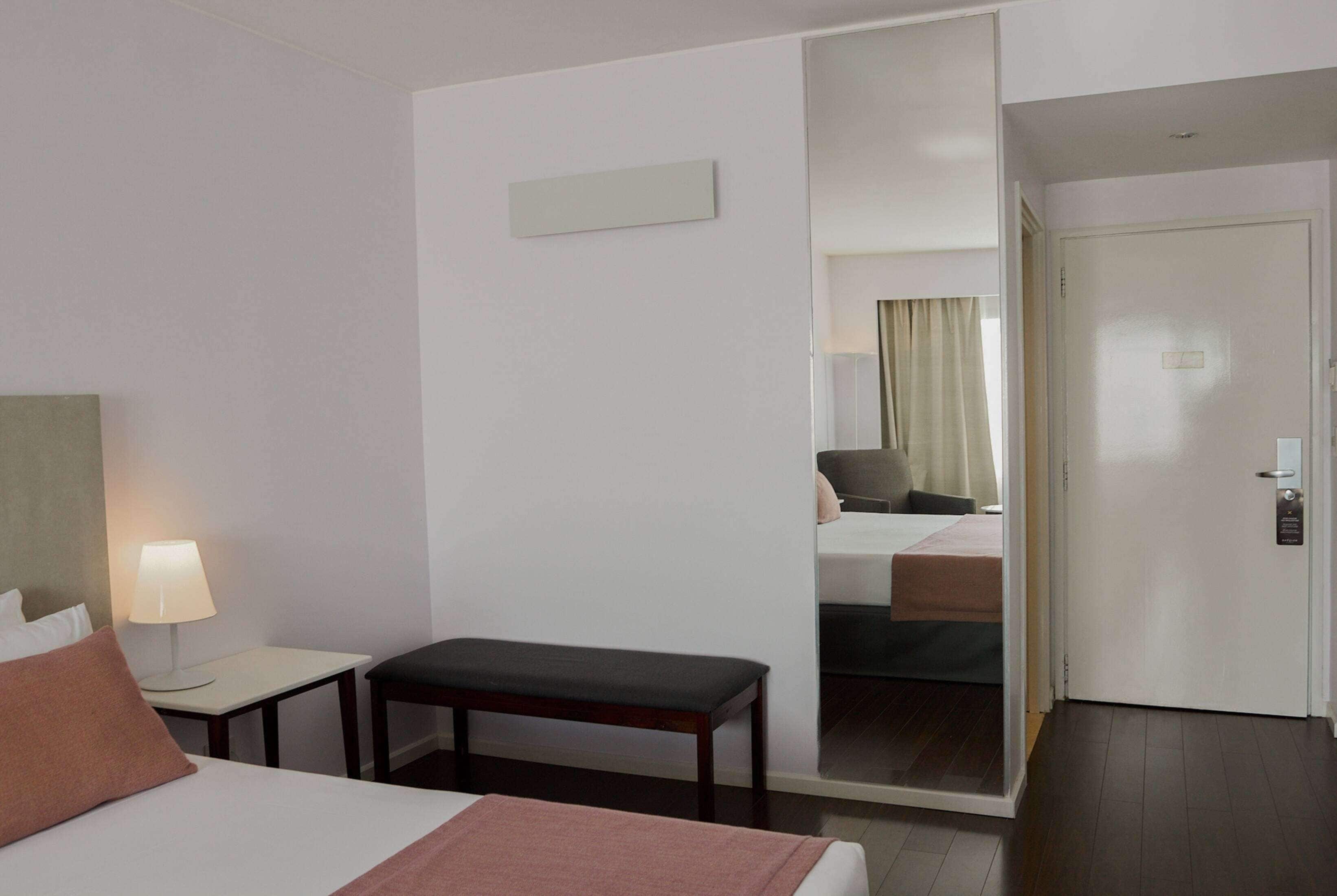 Una habitación de hotel con una cama, una mesita de noche con lámpara, un banco, un espejo y una puerta.