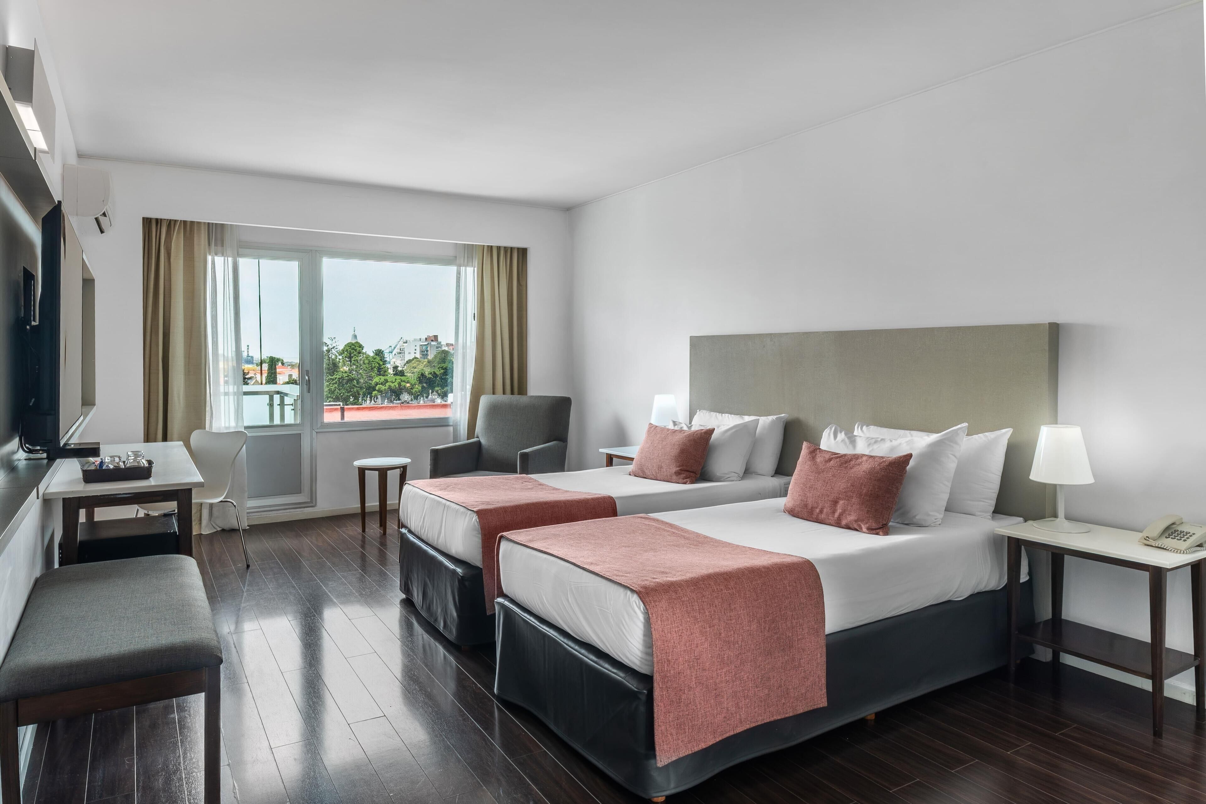 A hotel room with two beds, a desk, a chair, a bench, and a view of the city.