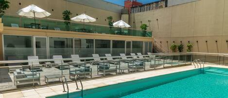 Una piscina en la azotea con sillones y sombrillas, barandilla de vidrio y una escalera que sube a un nivel superior.