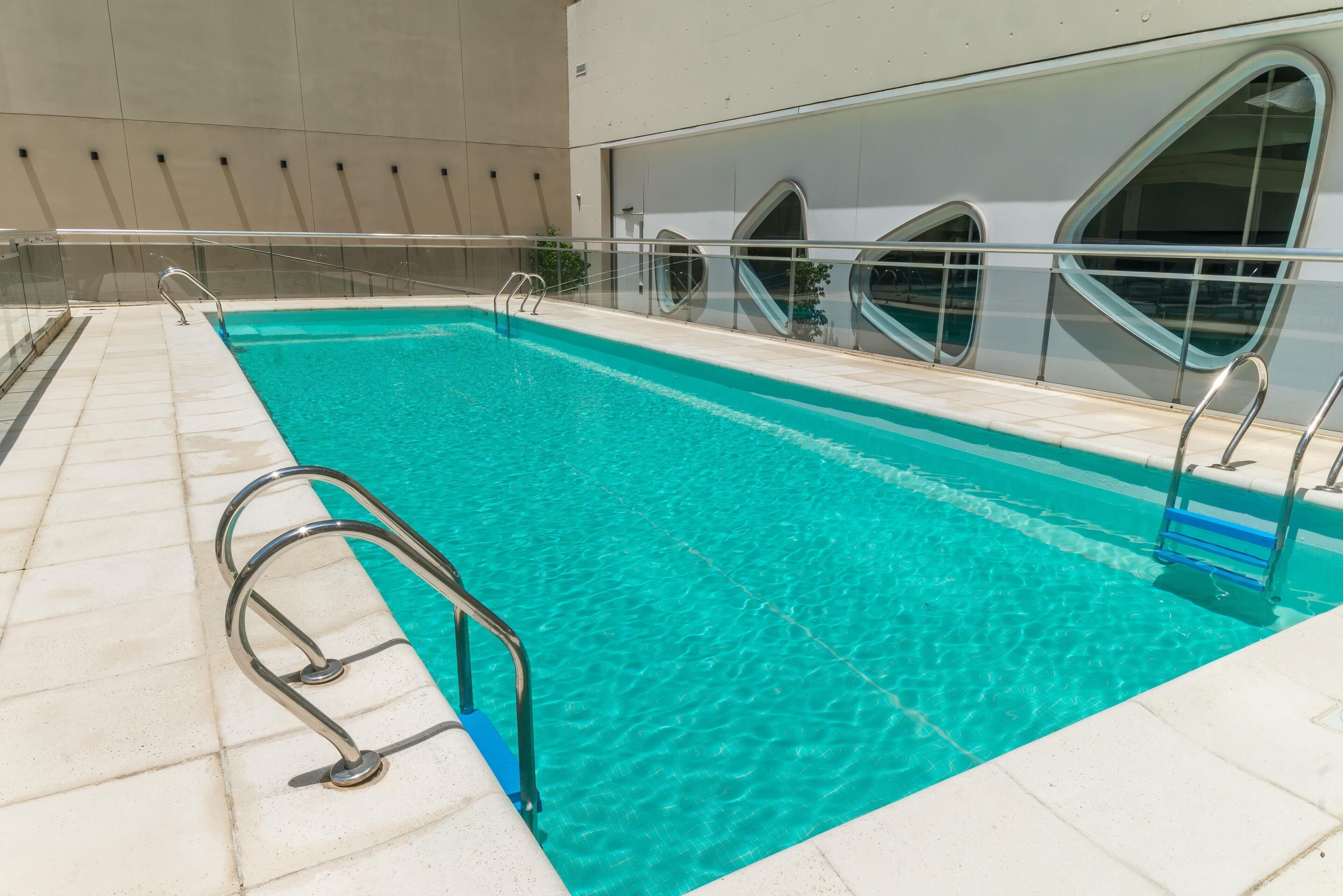Una piscina con agua cristalina, barandales de metal y un deck con baldosas.
