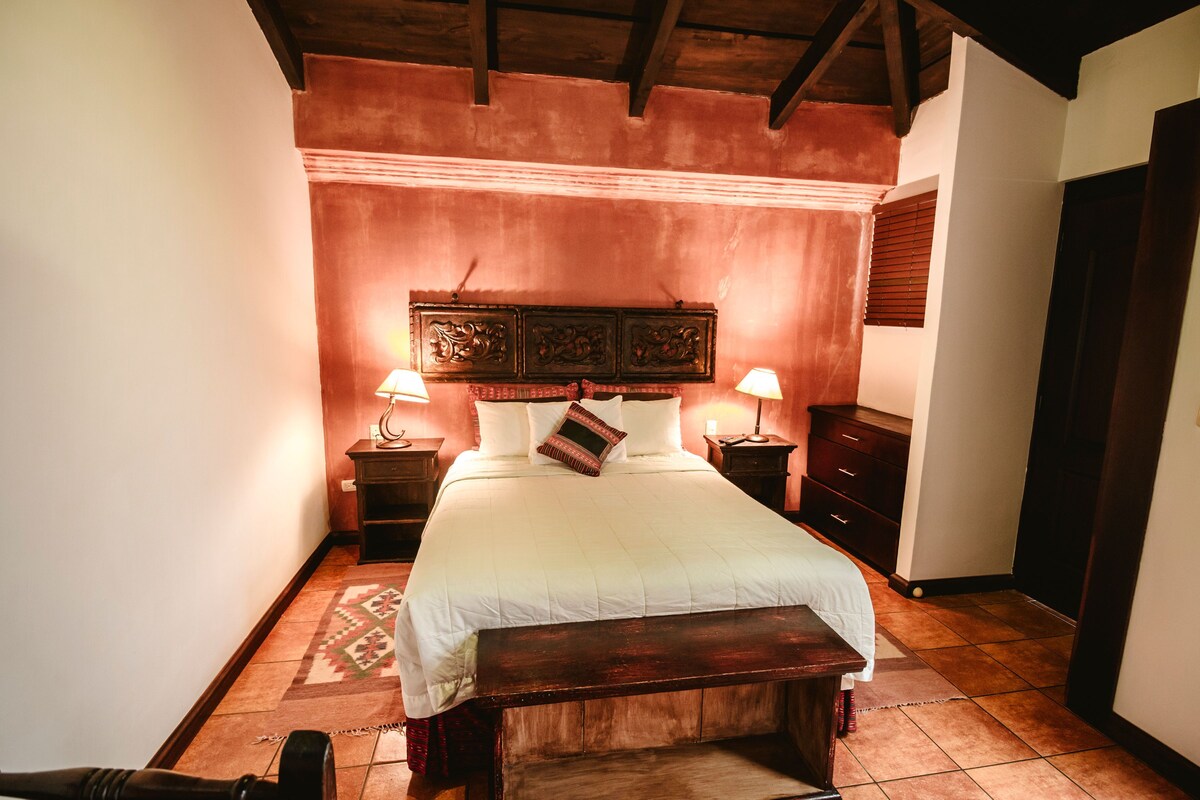 Un dormitorio con cabecera de madera, cama con ropa blanca, dos lámparas de noche, una mesita de noche, un banco de madera y un tapete estampado.