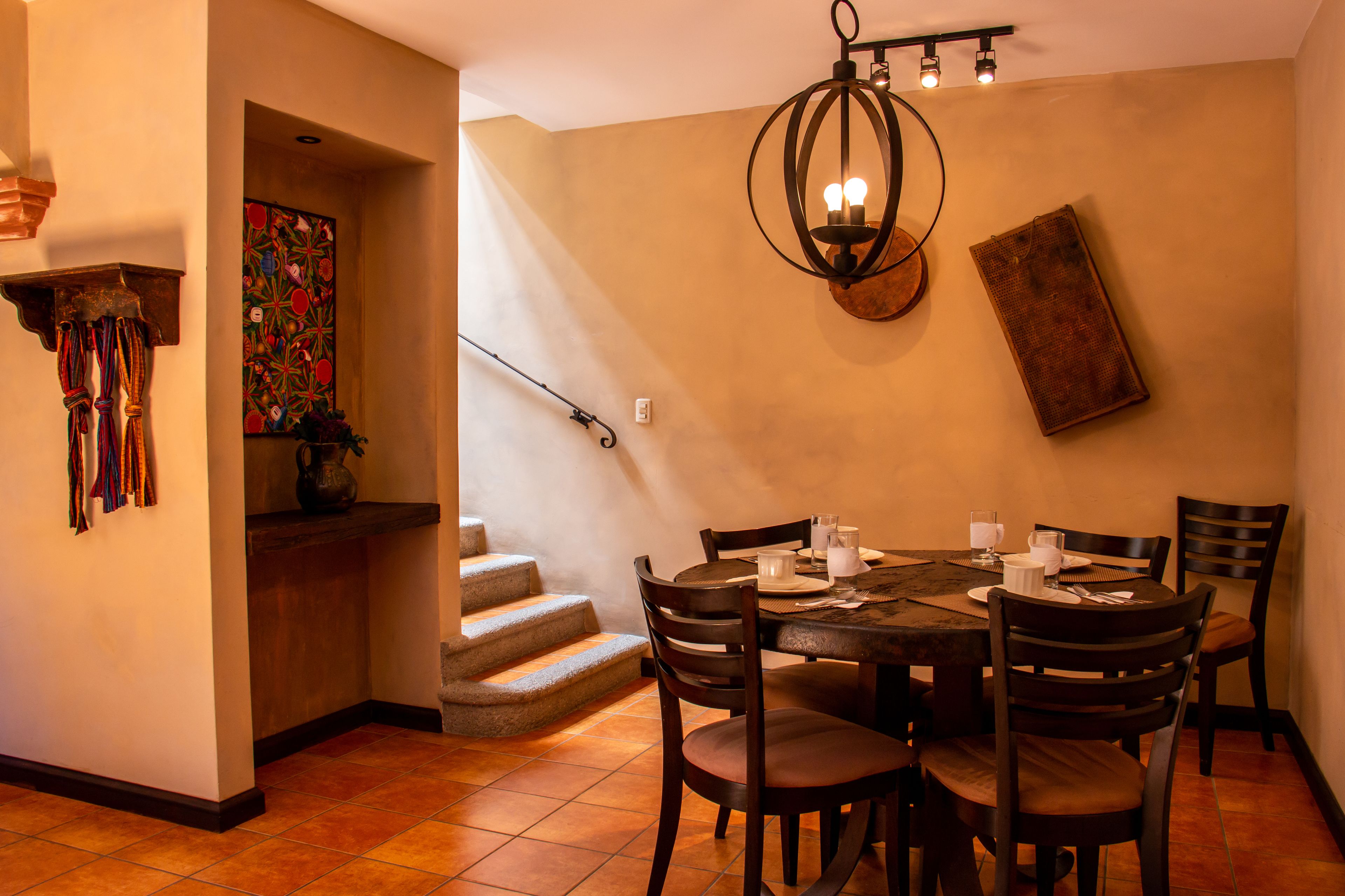 Un comedor con una mesa redonda, sillas y una araña. Una escalera conduce a otra habitación.
