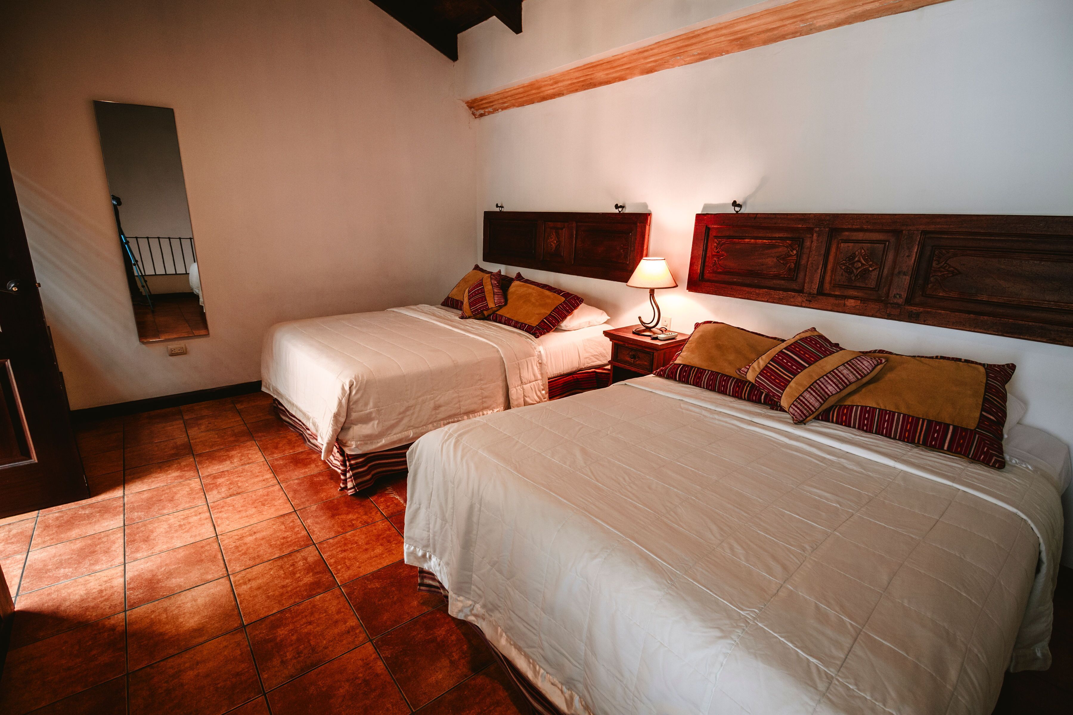 A bedroom with two beds, a nightstand with a lamp, a wooden headboard, and a mirror on the wall.