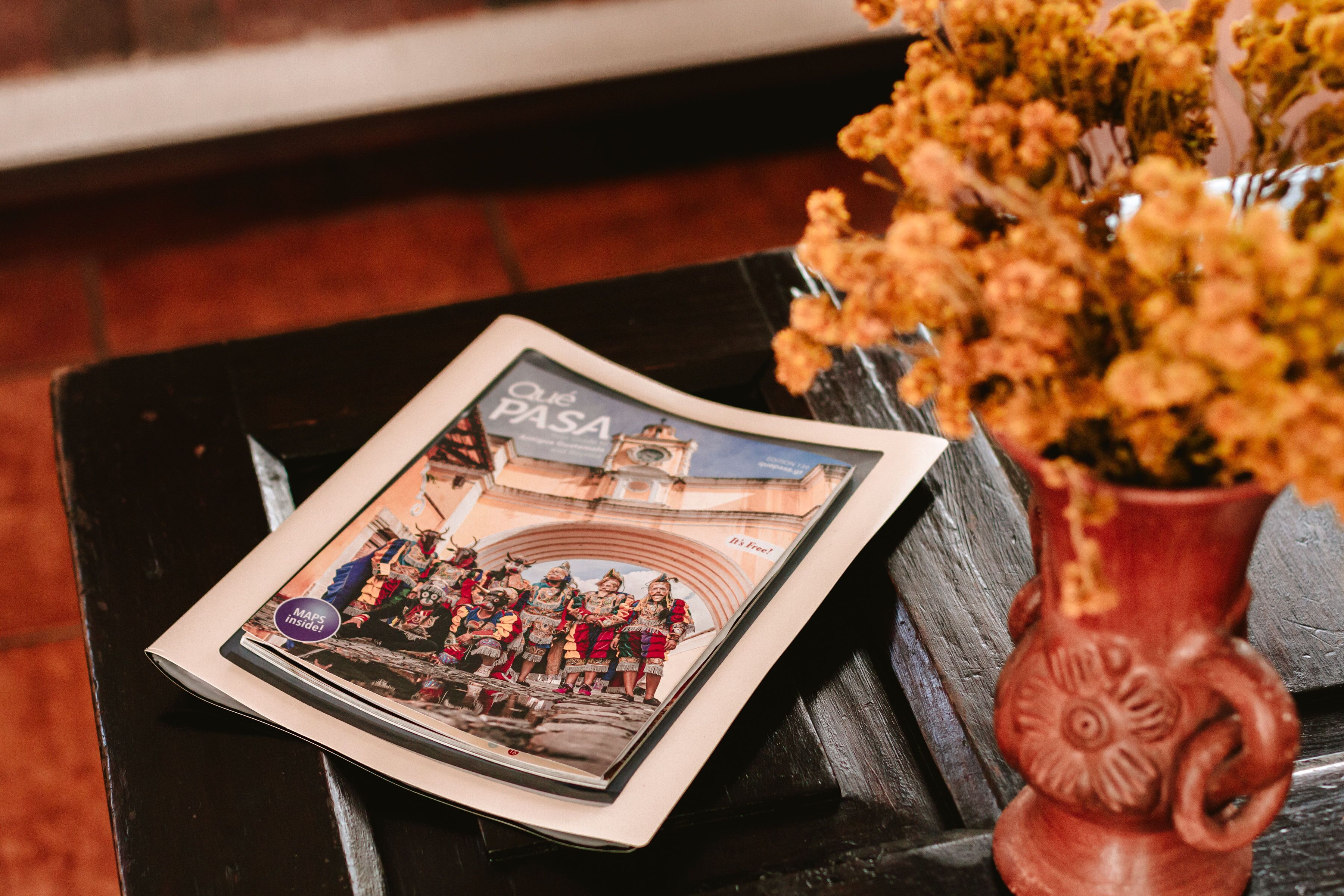 A magazine titled "Que PASA" on a black table next to a decorative vase with flowers.