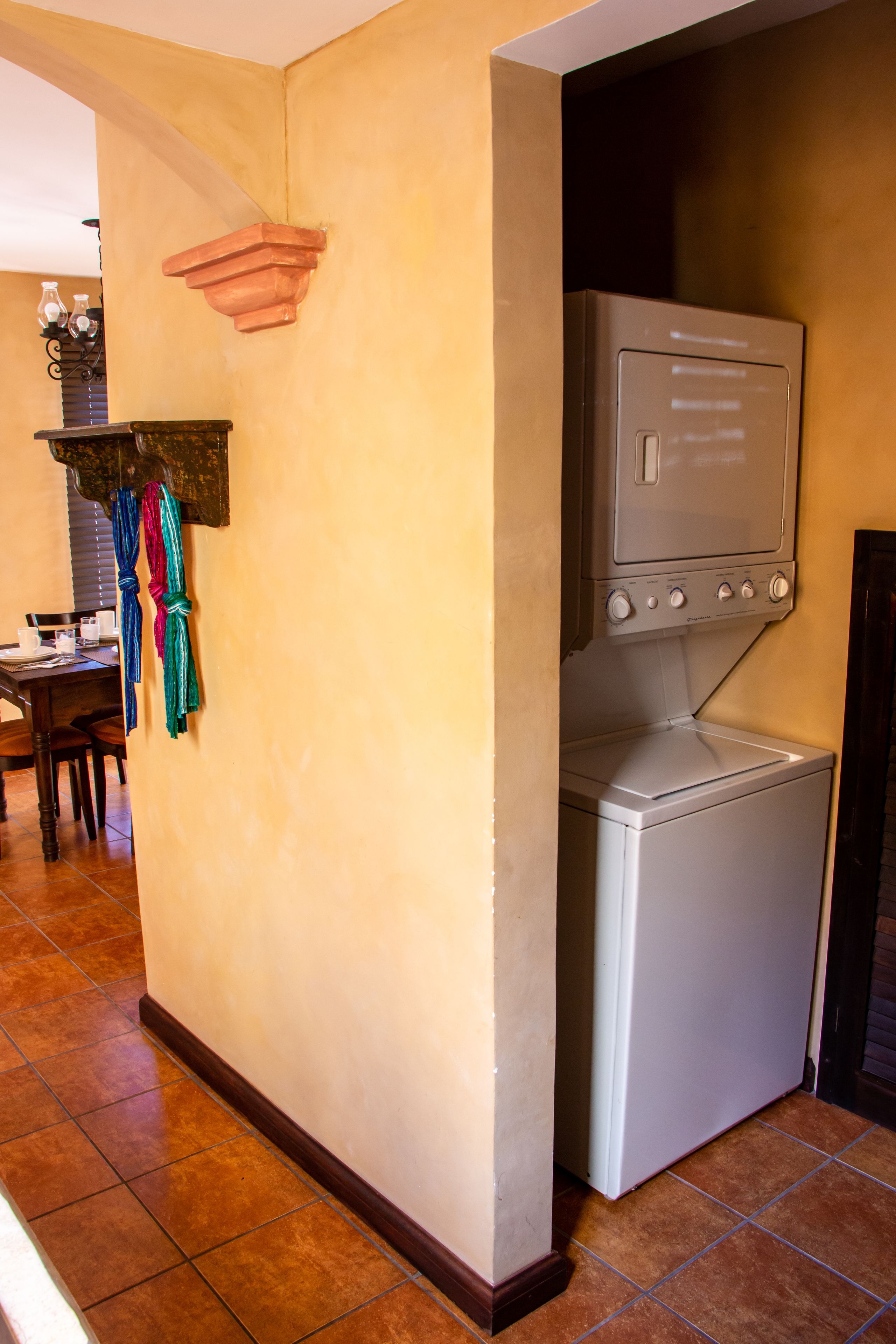 A laundry room with a washing machine and dryer, a wooden table with chairs, and a shelf with bottles and a clock.