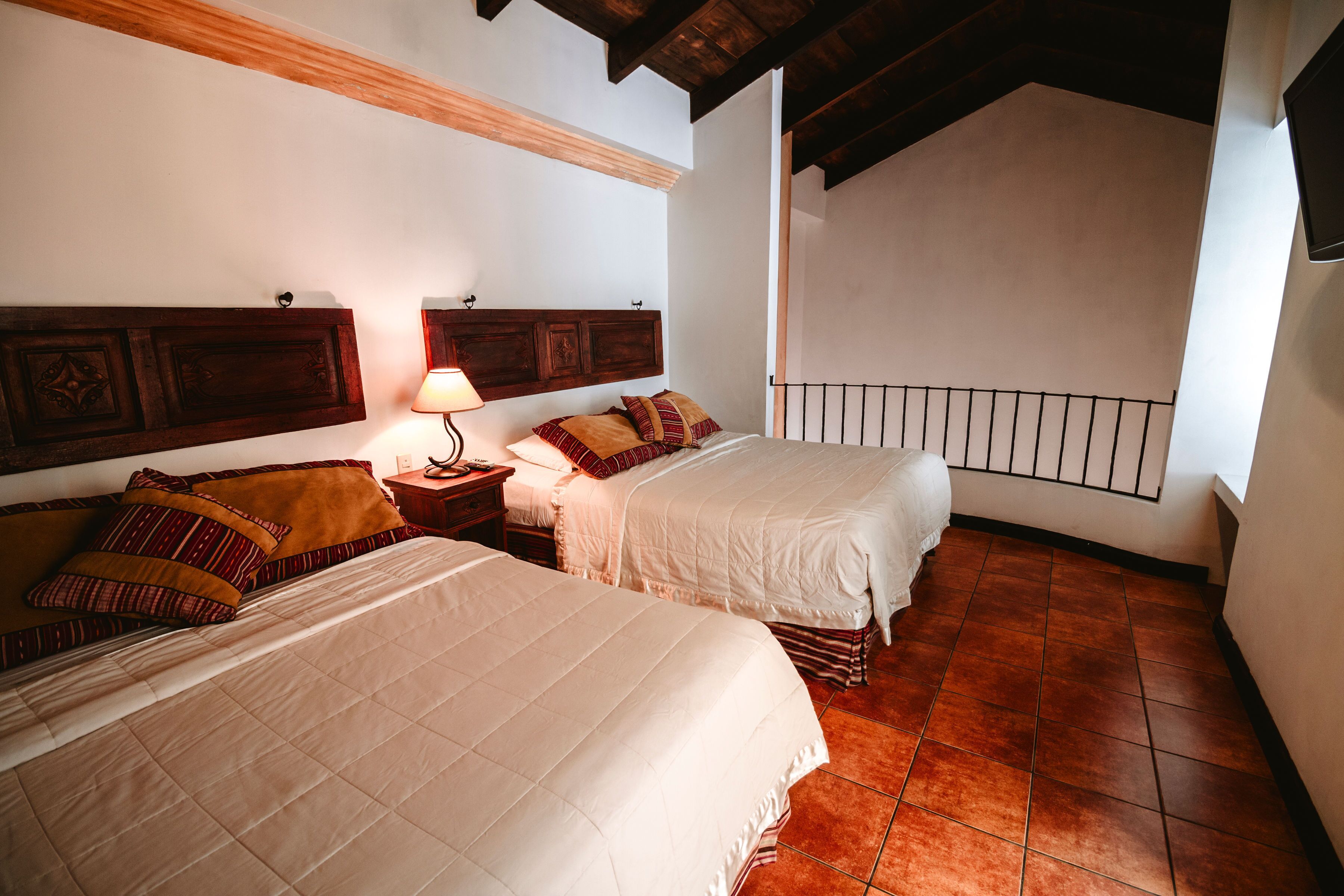A bedroom with two beds, wooden headboards, a nightstand with a lamp, and a tiled floor.