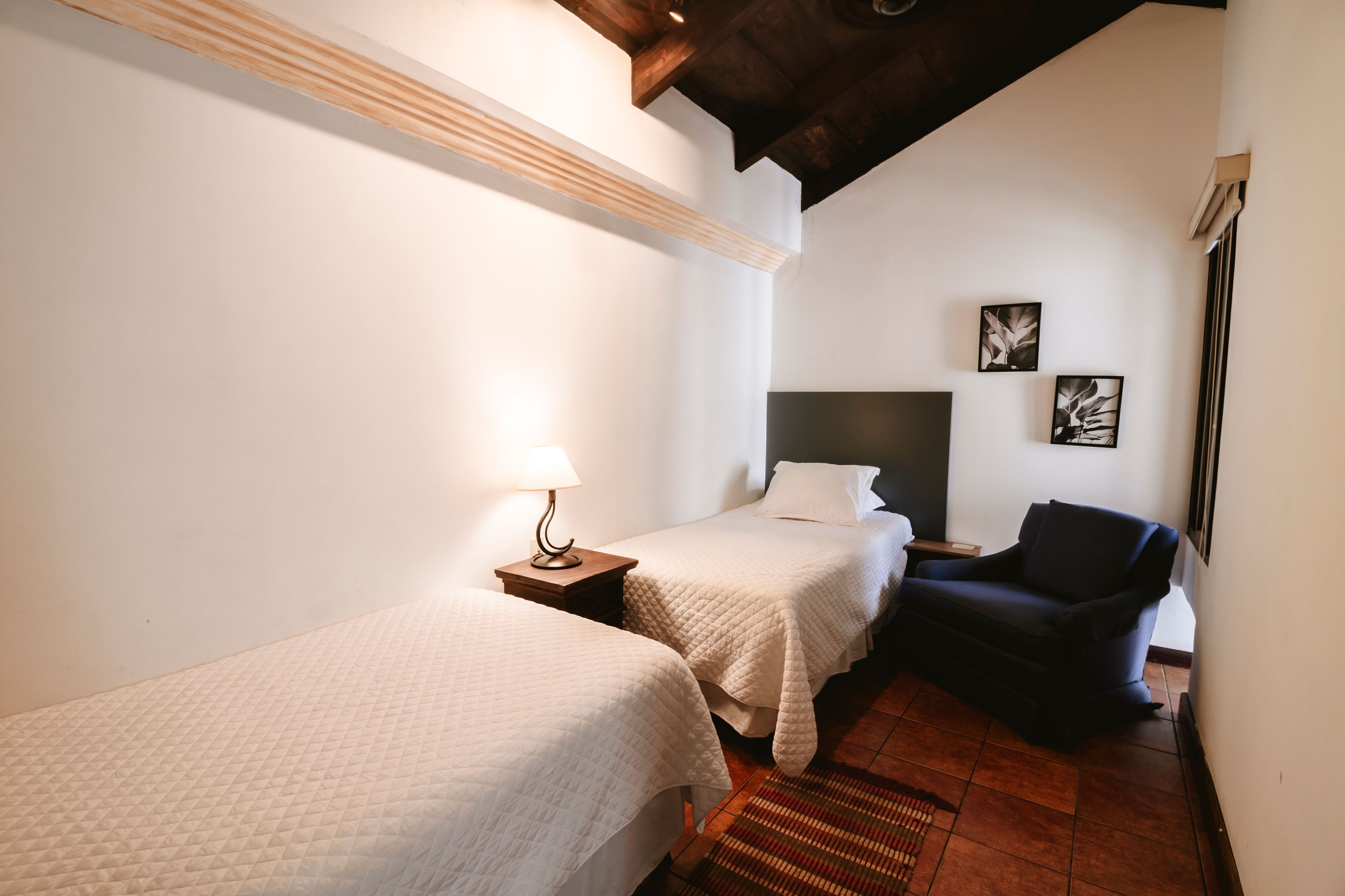 A hotel room with two single beds, a dark armchair, a small table with a lamp, and a wall-mounted TV.
