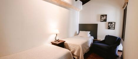 A hotel room with two single beds, a dark armchair, a small table with a lamp, and a wall-mounted TV.