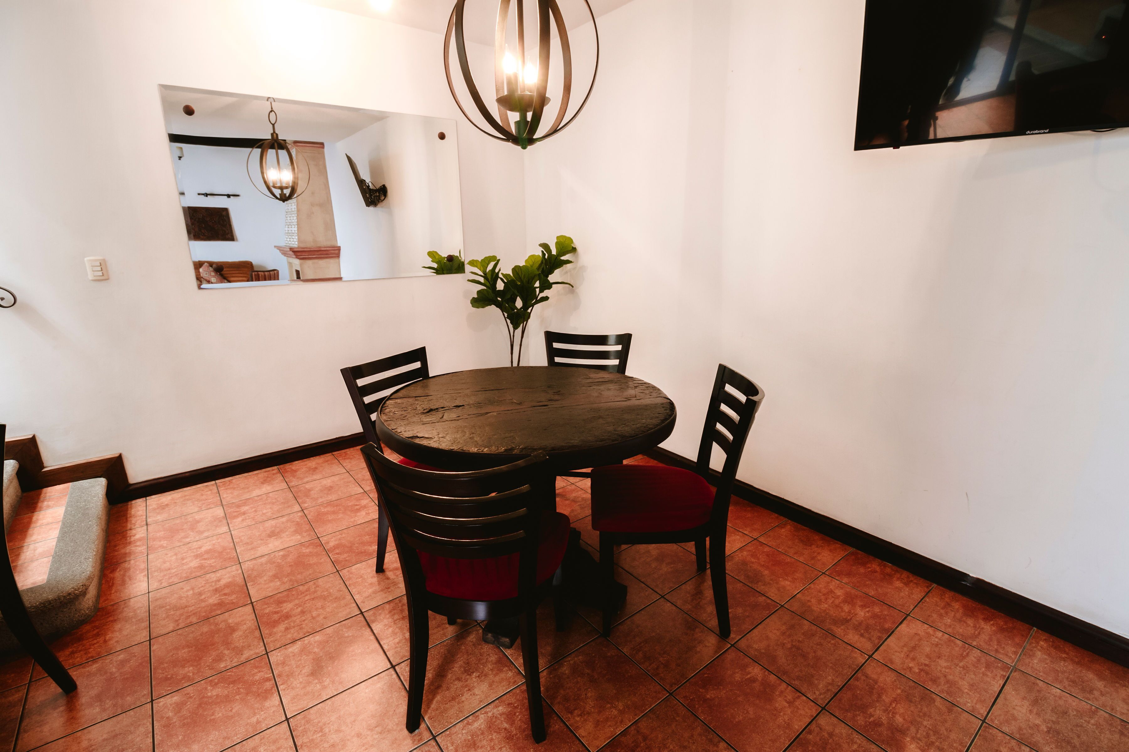 A dining area with a round table, four chairs, a mirror, a chandelier, and a potted plant.