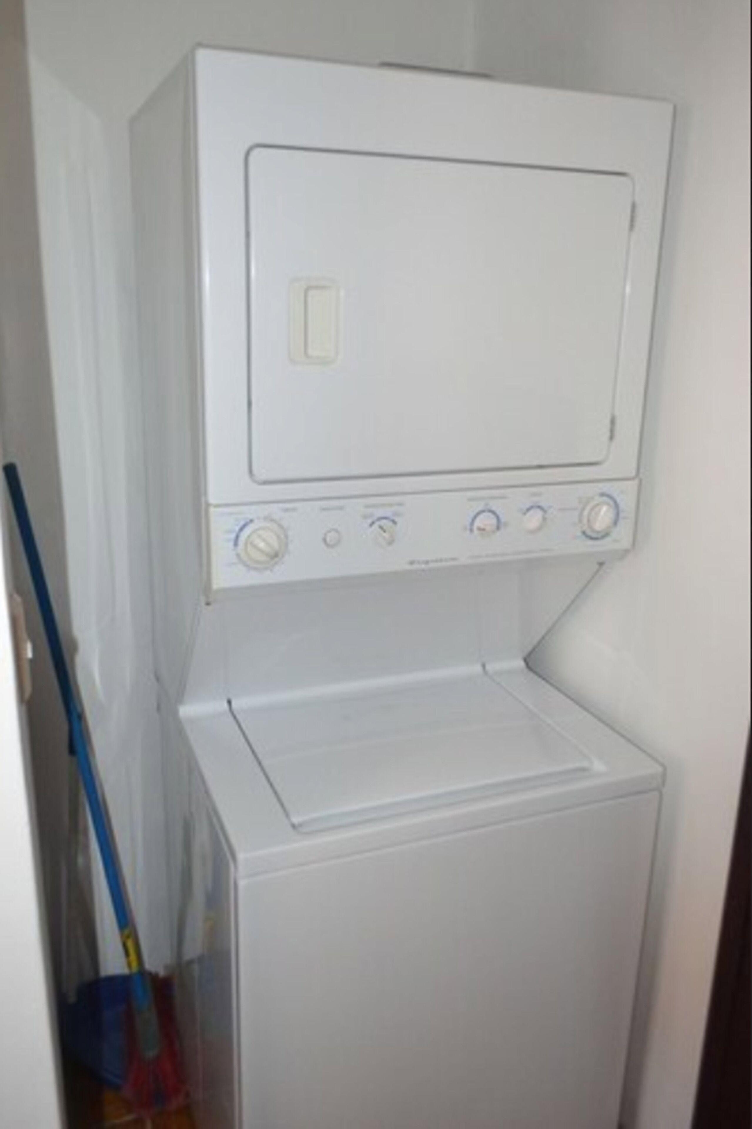 A white washing machine with a control panel, located in a laundry room with a mop leaning against the wall.