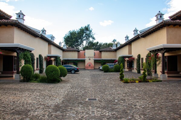 Un patio con un camino adoquinado, edificios de dos pisos con torres de campanas y automóviles estacionados.