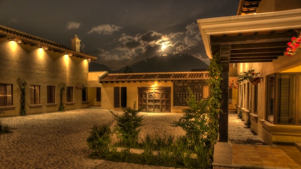 A courtyard with cobblestone flooring, a building with a tiled roof, and a mountain in the background.