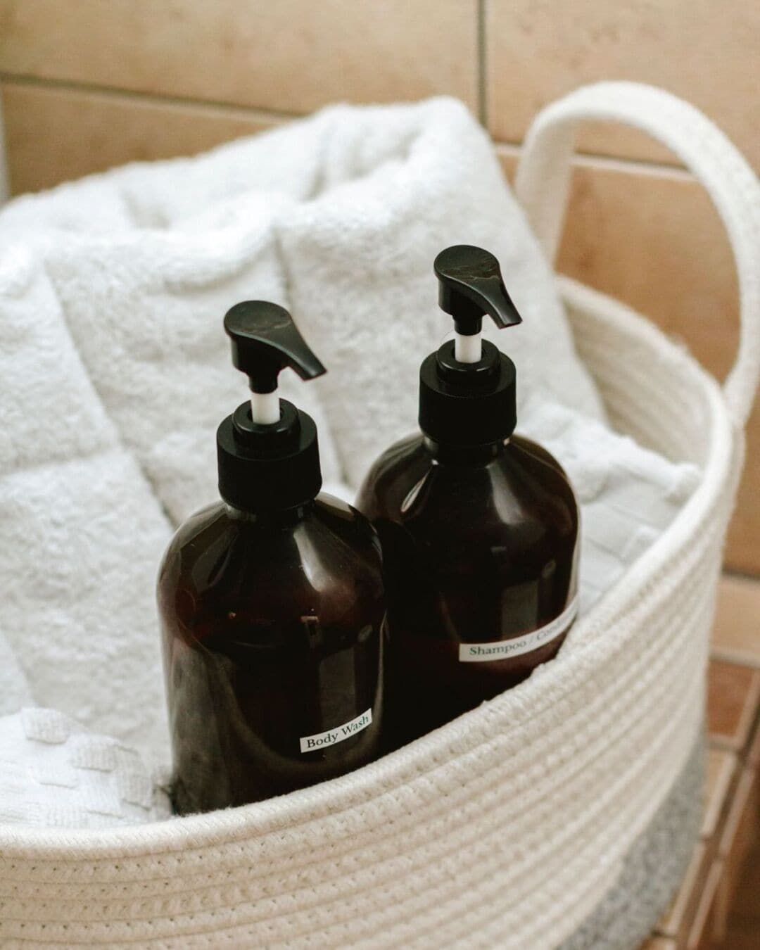 A white woven basket containing two dark brown bottles with pump dispensers, labeled 'Body Wash' and 'Shampoo / Conditioner'.