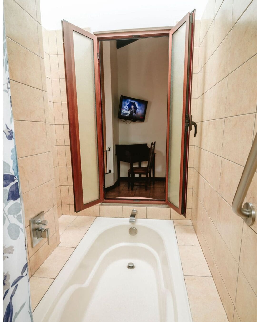 A bathroom with a bathtub, a television mounted on the wall, and a chair visible through an open door.