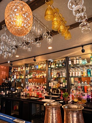 Zona de bar con estantes llenos de botellas de licores, una jarra de cobre y copas colgantes.