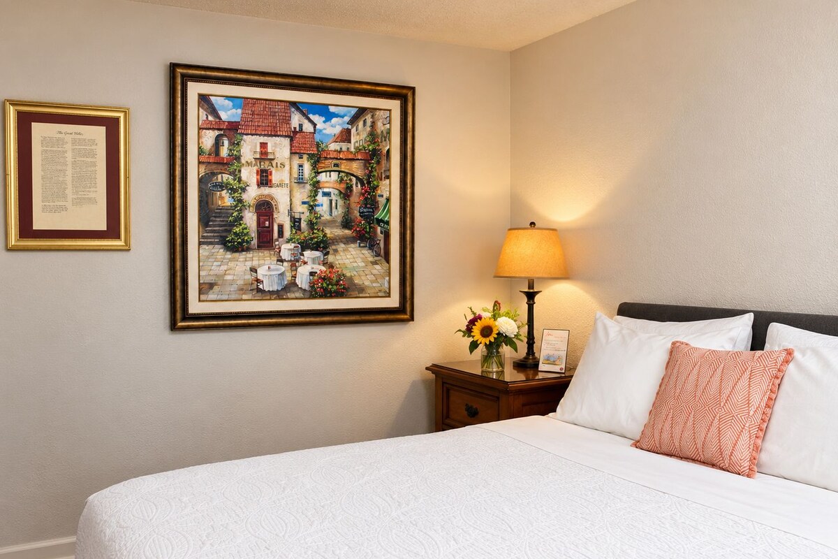 Habitación de hotel con cama, mesita de noche con lámpara y flores, un cuadro enmarcado y un certificado en la pared.