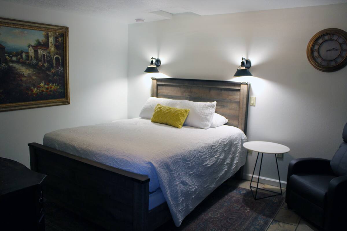 Una cama bien hecha con cabecera de madera, una mesita redonda pequeña, una silla y un cuadro en la pared.