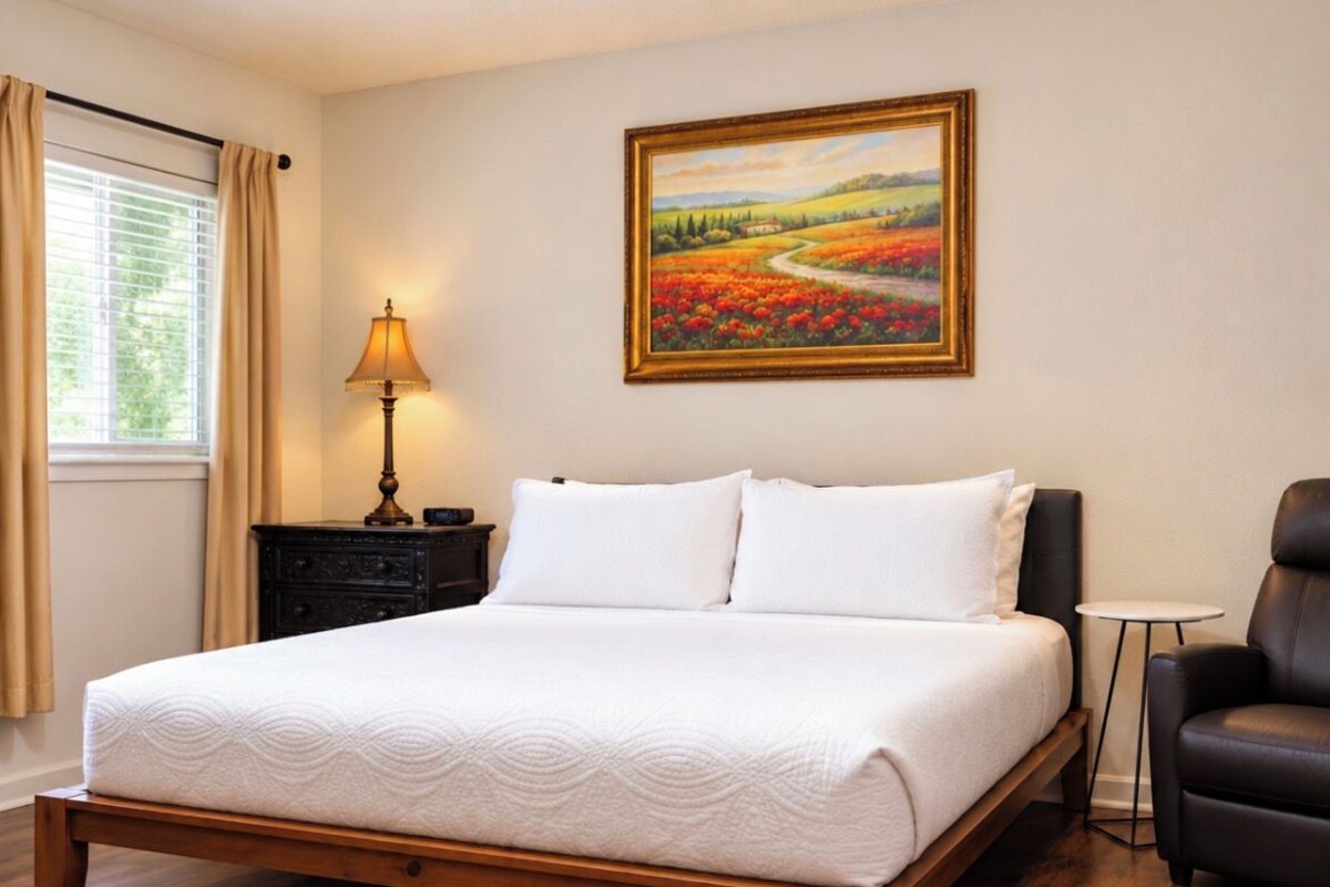 Una cama bien hecha con edredón blanco y almohadas, una mesita de noche con una lámpara, un cuadro de paisaje enmarcado en la pared y un sillón de cuero.