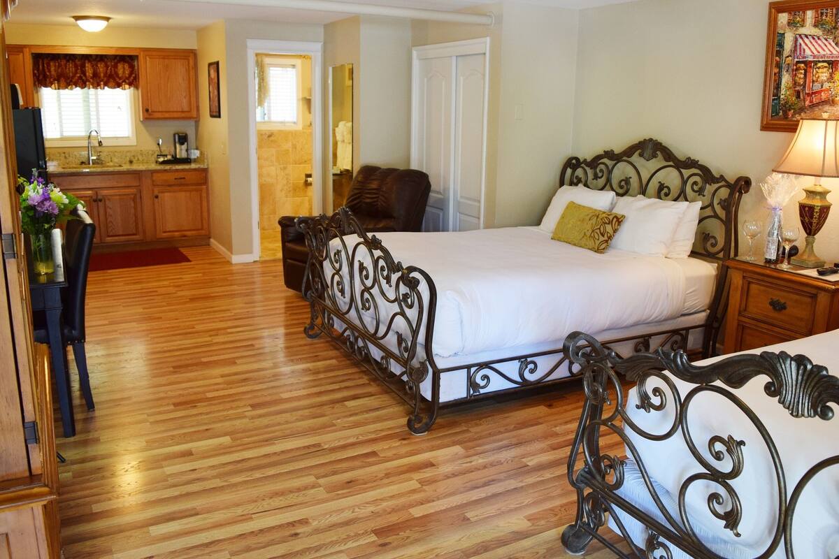 Habitación de hotel con piso de madera, cama con estructura metálica, mesita de noche con lámpara y vistas a la cocina y al baño.