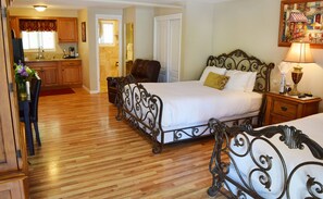 A hotel room with a wooden floor, a bed with a metal frame, a bedside table with a lamp, and a view of the kitchen and bathroom.