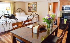 Habitación de hotel con cama, una mesa de comedor con sillas, un florero con flores y una zona de cocina con estufa y horno.