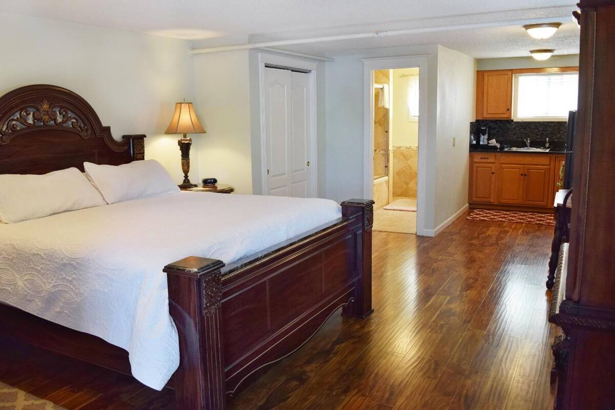 Un dormitorio con una cama grande, una mesita de noche, una lámpara y un cabecero de madera. Se ve una puerta que da a un baño y una zona de cocina.