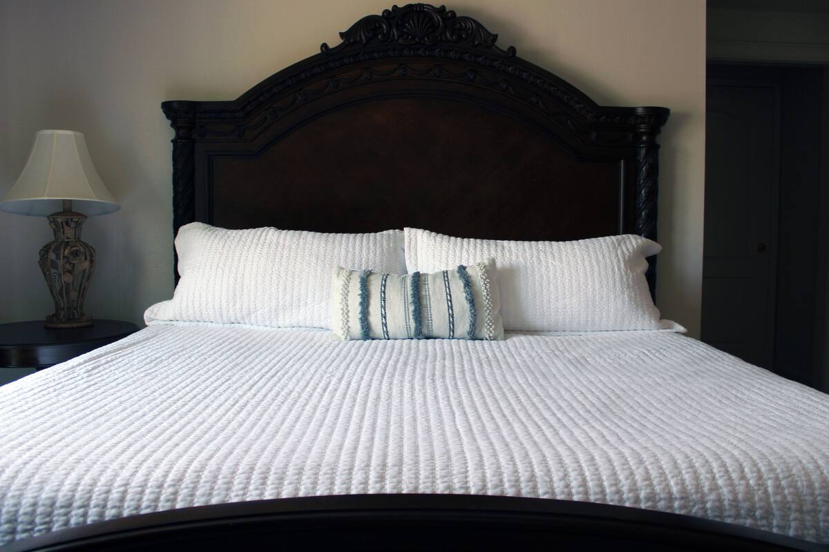 Una cama bien hecha con un edredón blanco y dos almohadas, un cabecero de madera oscura y una mesita de noche con una lámpara.