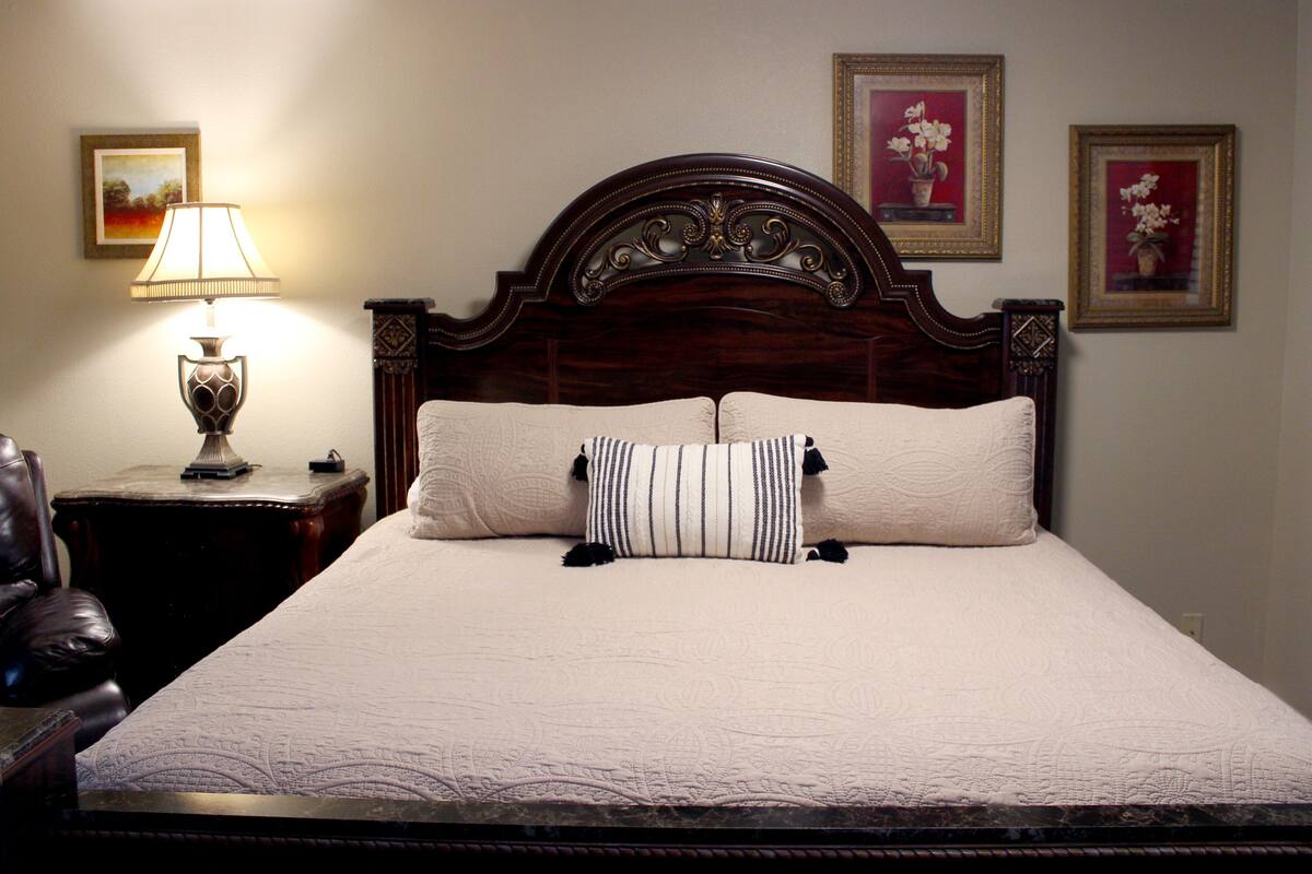 Una cama bien hecha con cabecera de madera, lámpara de noche y cuadros en la pared.