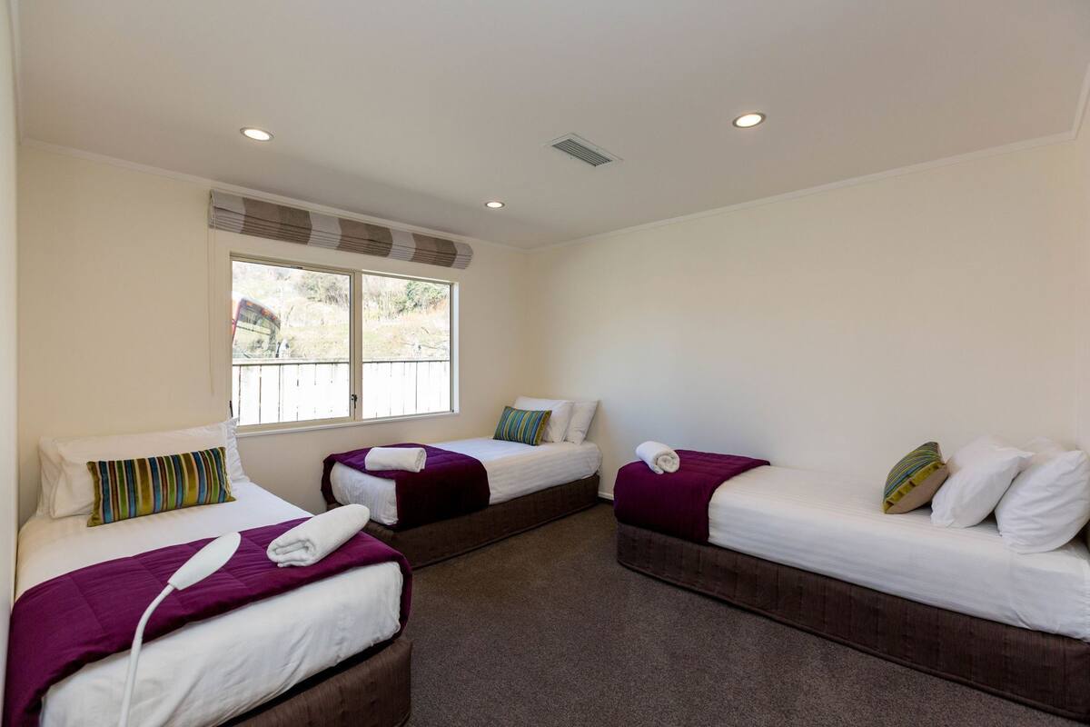 A room with three beds, each with white bedding and purple accents, a large window with a view of greenery, and a ceiling with recessed lighting.