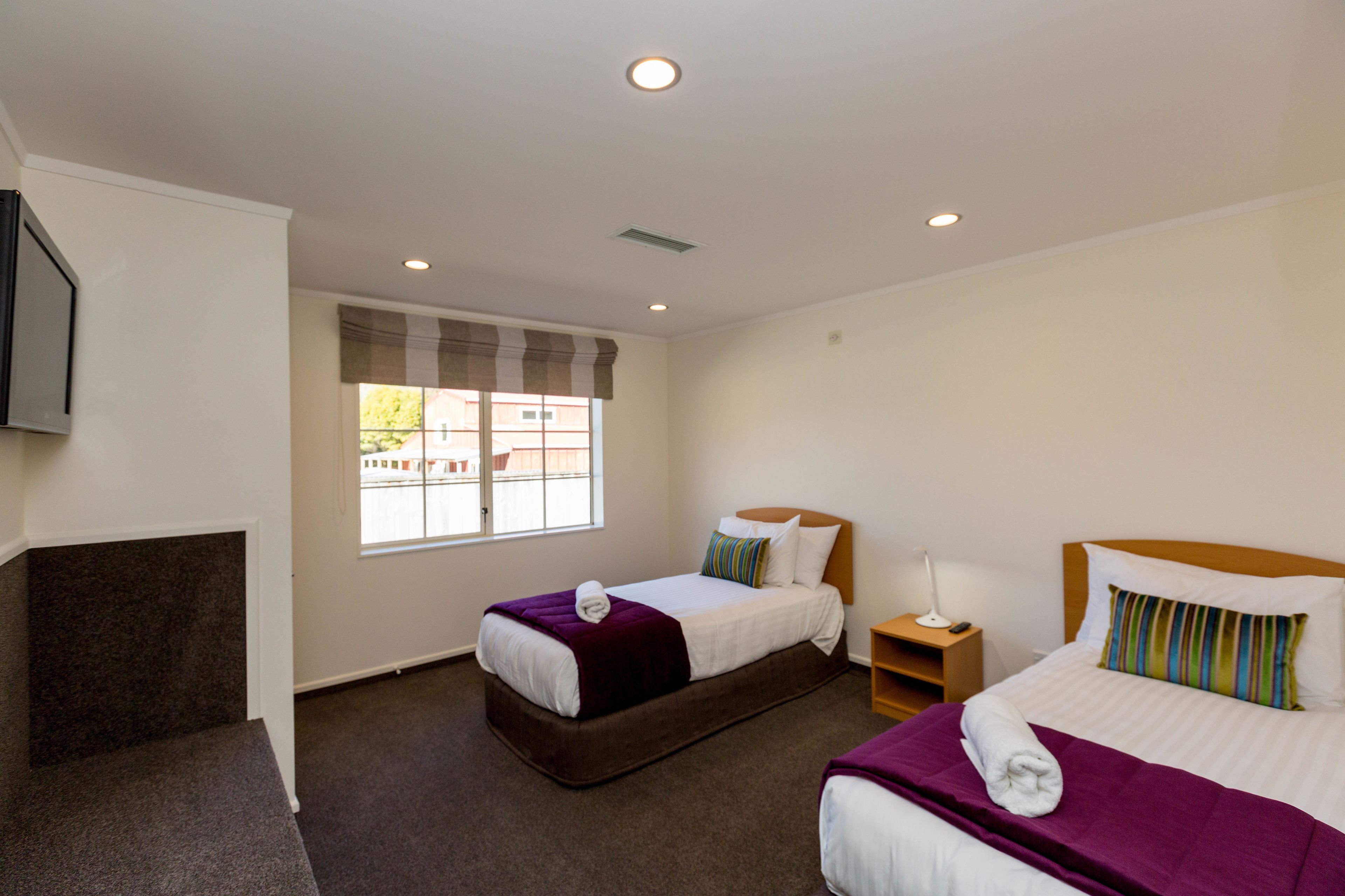 A hotel room with two beds, a TV mounted on the wall, a window with blinds, and a small bedside table with a lamp.