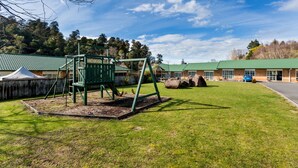 A playground with swings and a climbing structure, a building with a green roof, and a parking area. - Aspen Court Motel Taihape (Taihape)