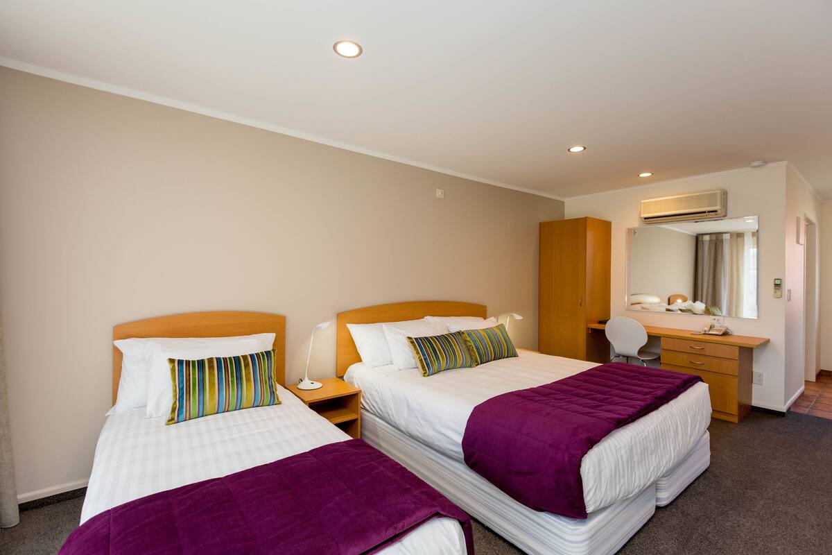 A hotel room with two beds, a desk, a chair, a wardrobe, and an air conditioning unit.
