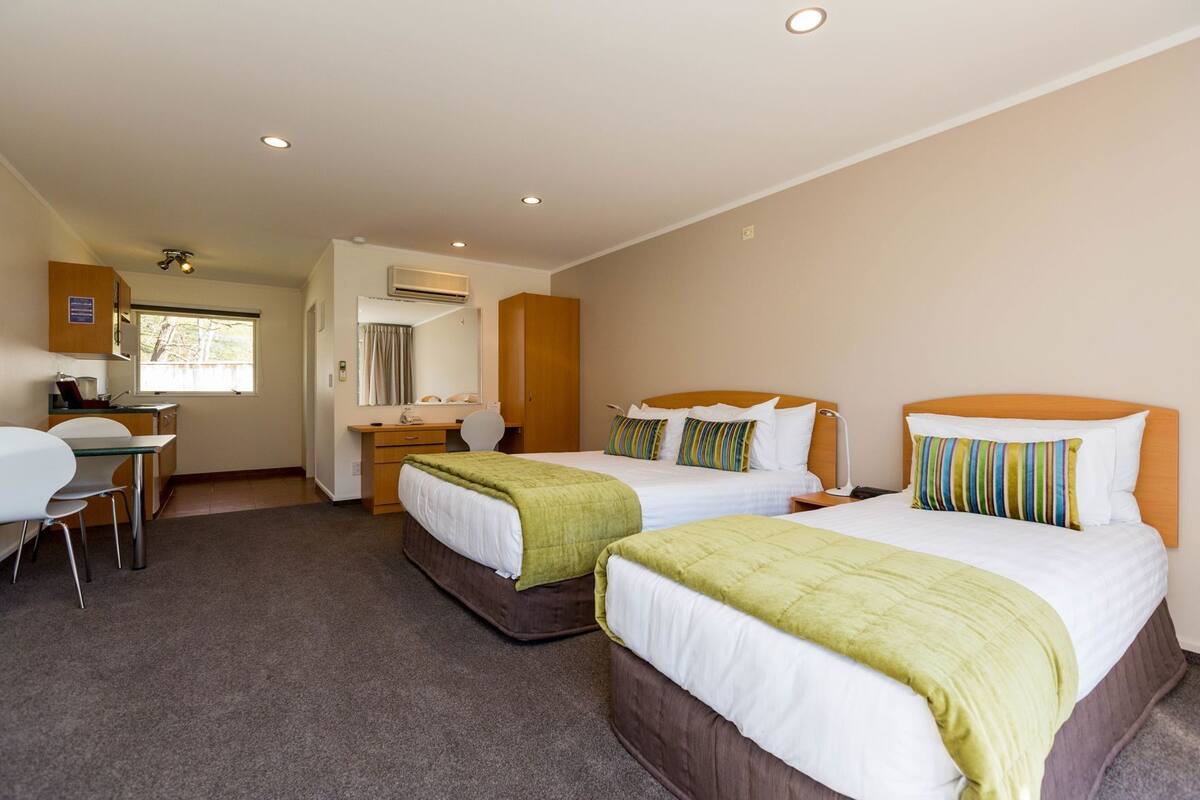 A hotel room with two beds, a desk, a chair, an air conditioning unit, and a window.