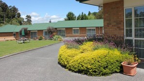 A building with a green roof, brick walls, and large windows. There is a paved driveway and a landscaped area with yellow and purple plants. - Aspen Court Motel Taihape (Taihape)