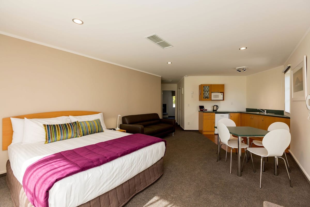 A hotel room with a bed, a sofa, a dining table with chairs, a kitchenette, and a bathroom visible in the background.