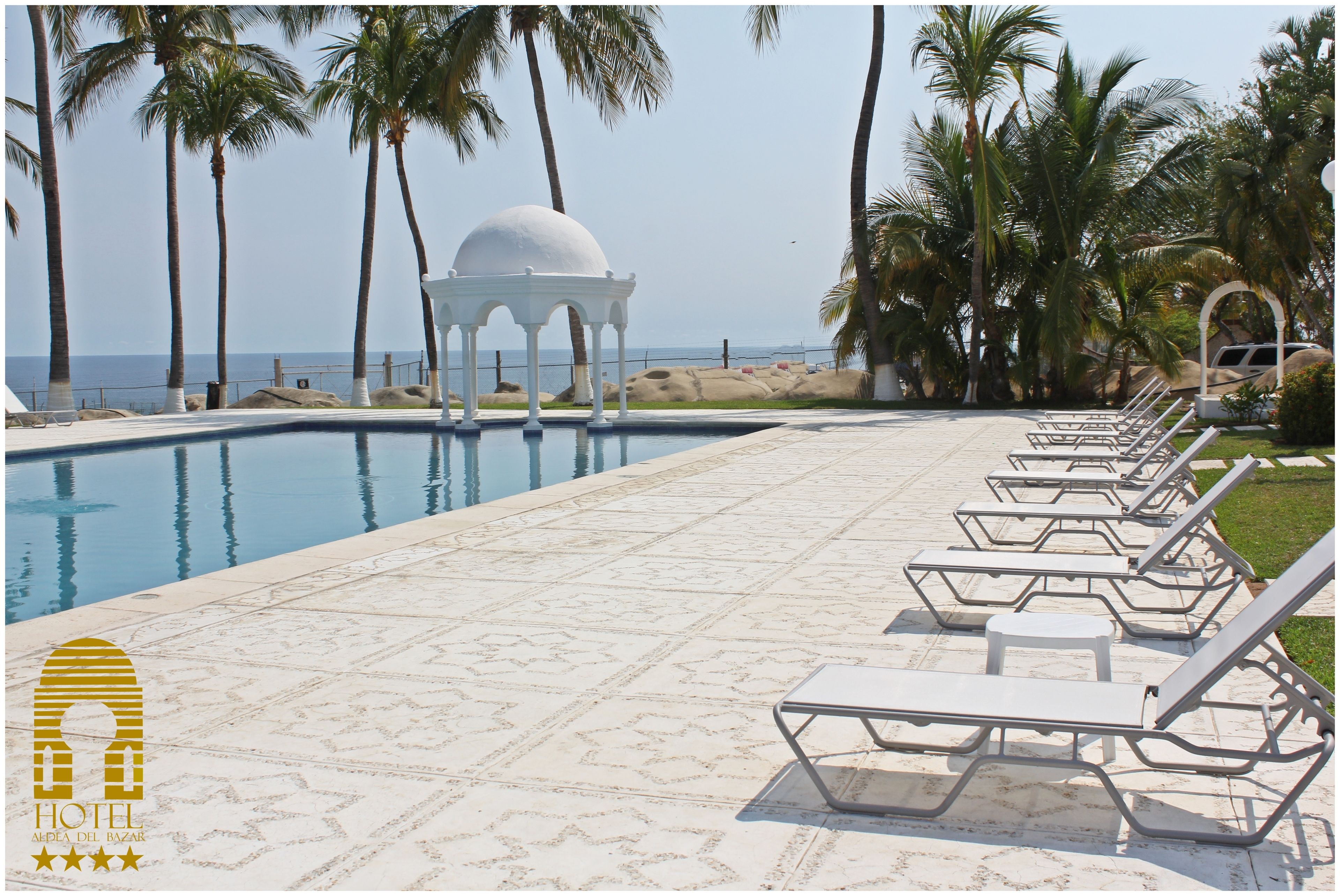 Área ao lado da piscina com espreguiçadeiras, um gazebo branco e palmeiras.