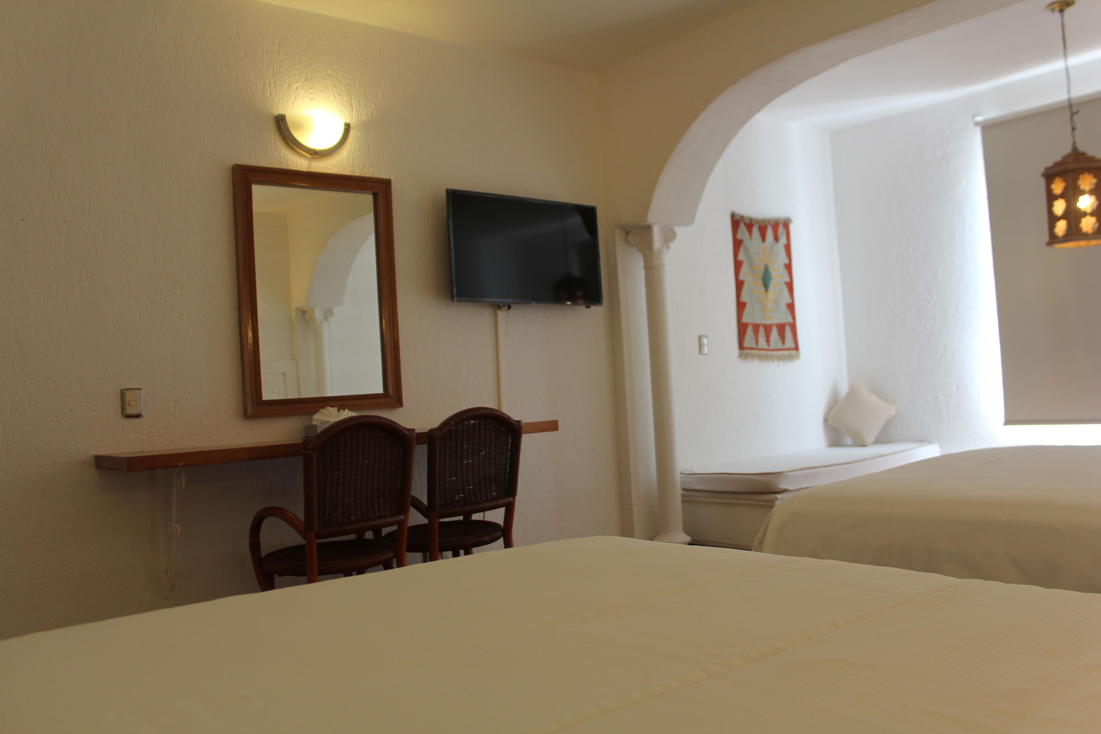 A hotel room with two beds, a TV mounted on the wall, a desk with chairs, and a decorative wall hanging.