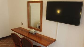 A room with a wooden desk, two wicker chairs, a mirror, and a mounted TV.