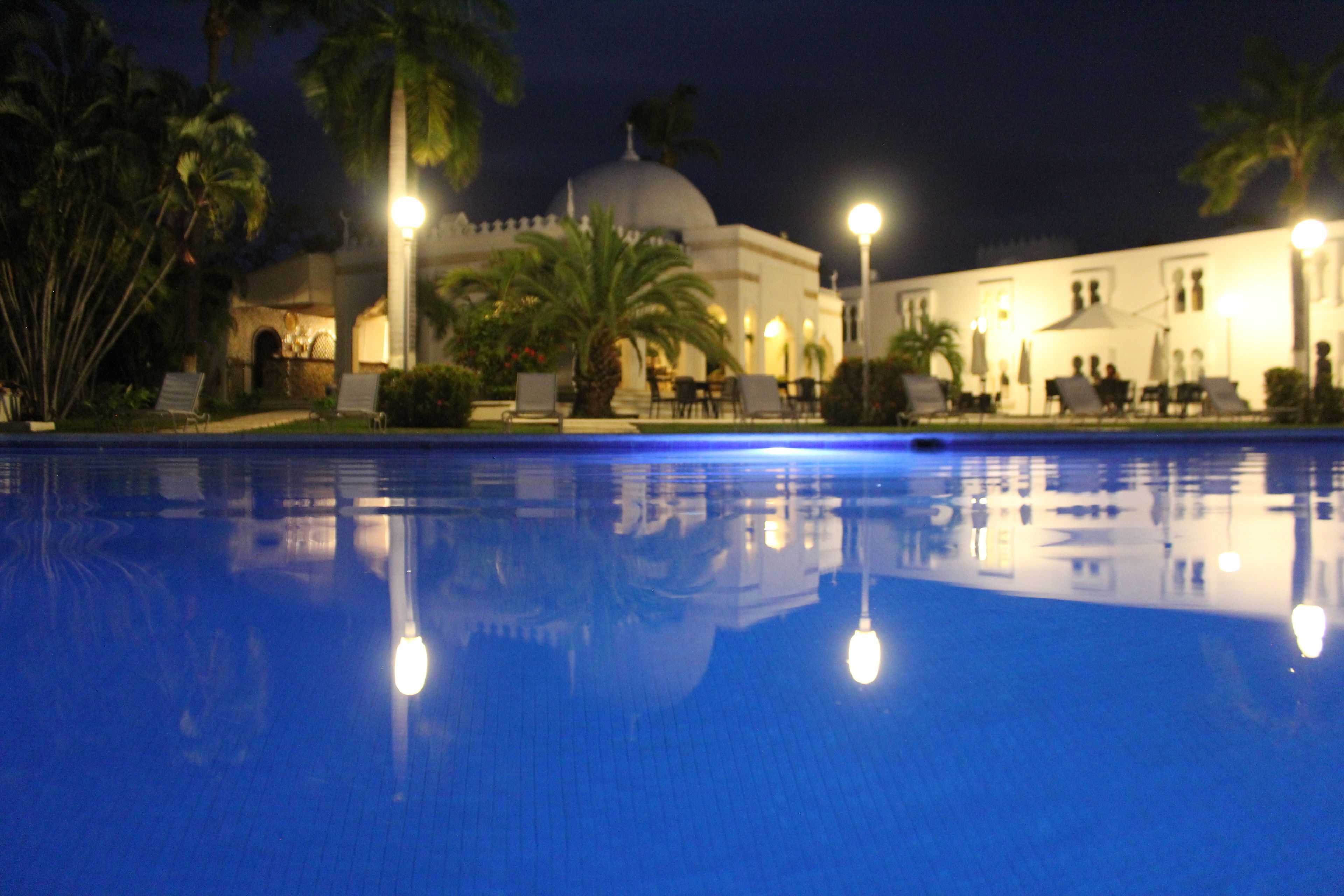 A nighttime view of a swimming pool with reflections of palm trees and a building with a dome.