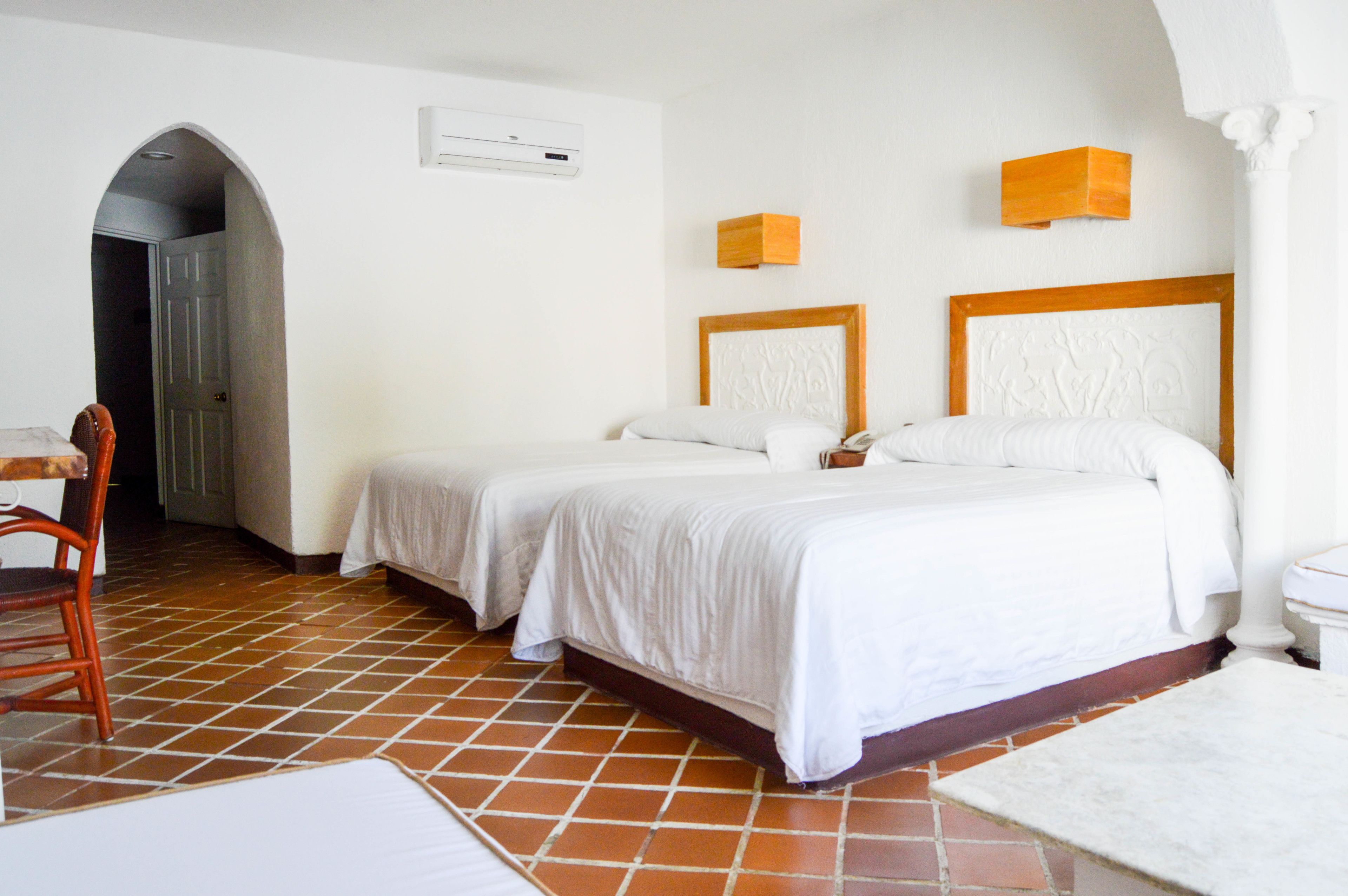 A hotel room with two beds, a chair, a desk, and a tiled floor.