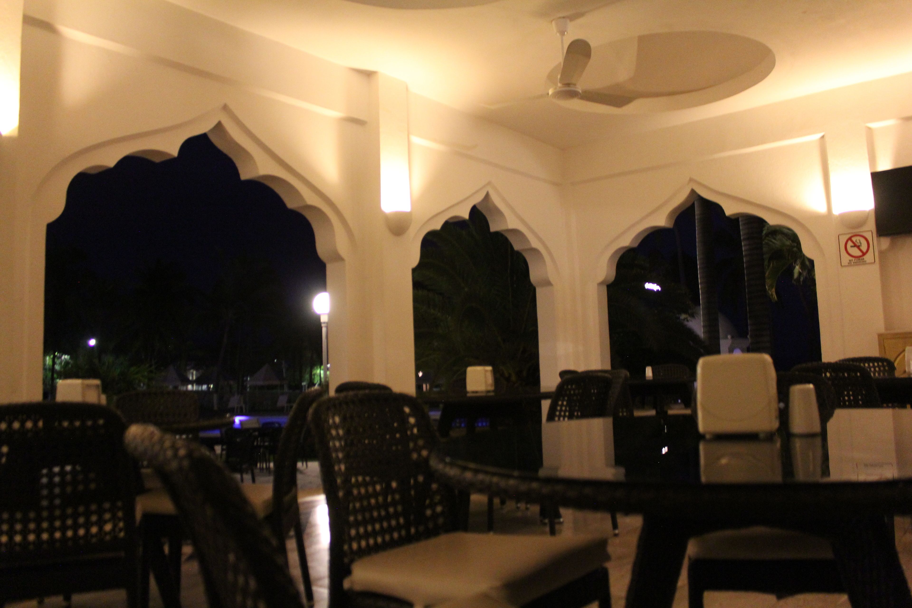 An indoor dining area with high-backed chairs and tables, large decorative arches, and a ceiling fan.
