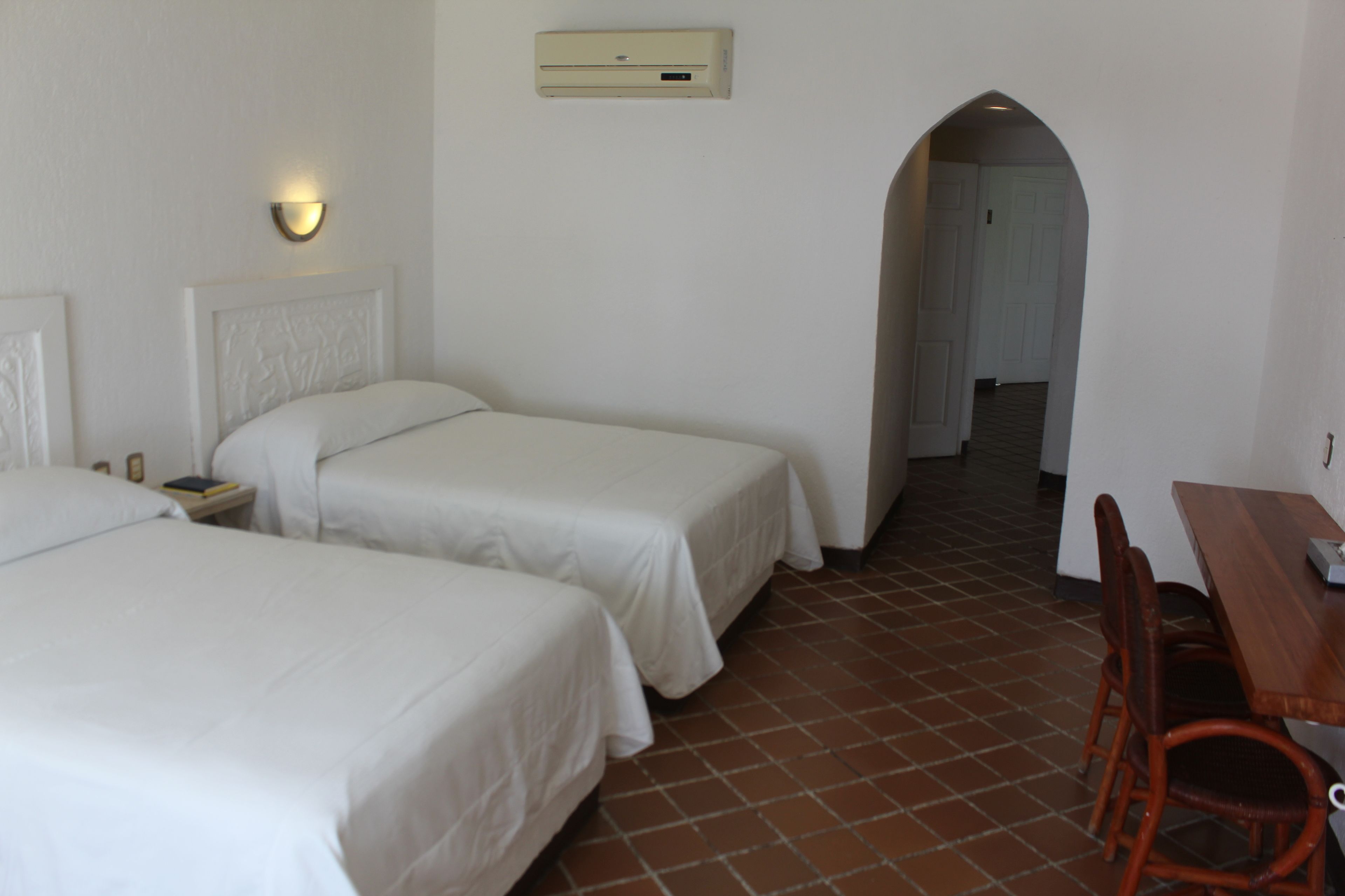 Habitación de hotel con dos camas individuales, una mesa de madera con sillas, aire acondicionado de pared y un pasillo que conduce a otra habitación.