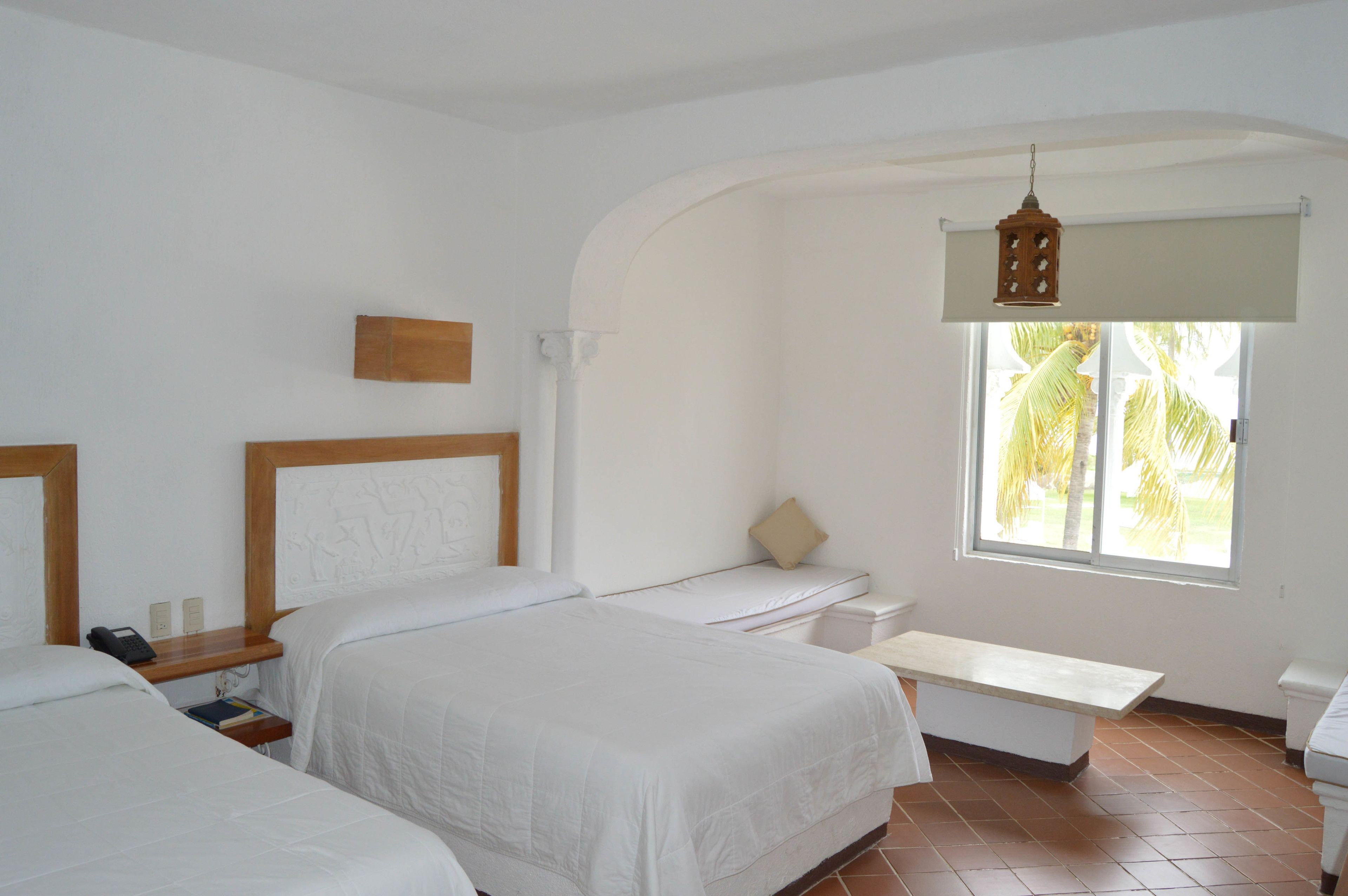 A room with two beds, a small table, a bench, and a window with a view of palm trees.